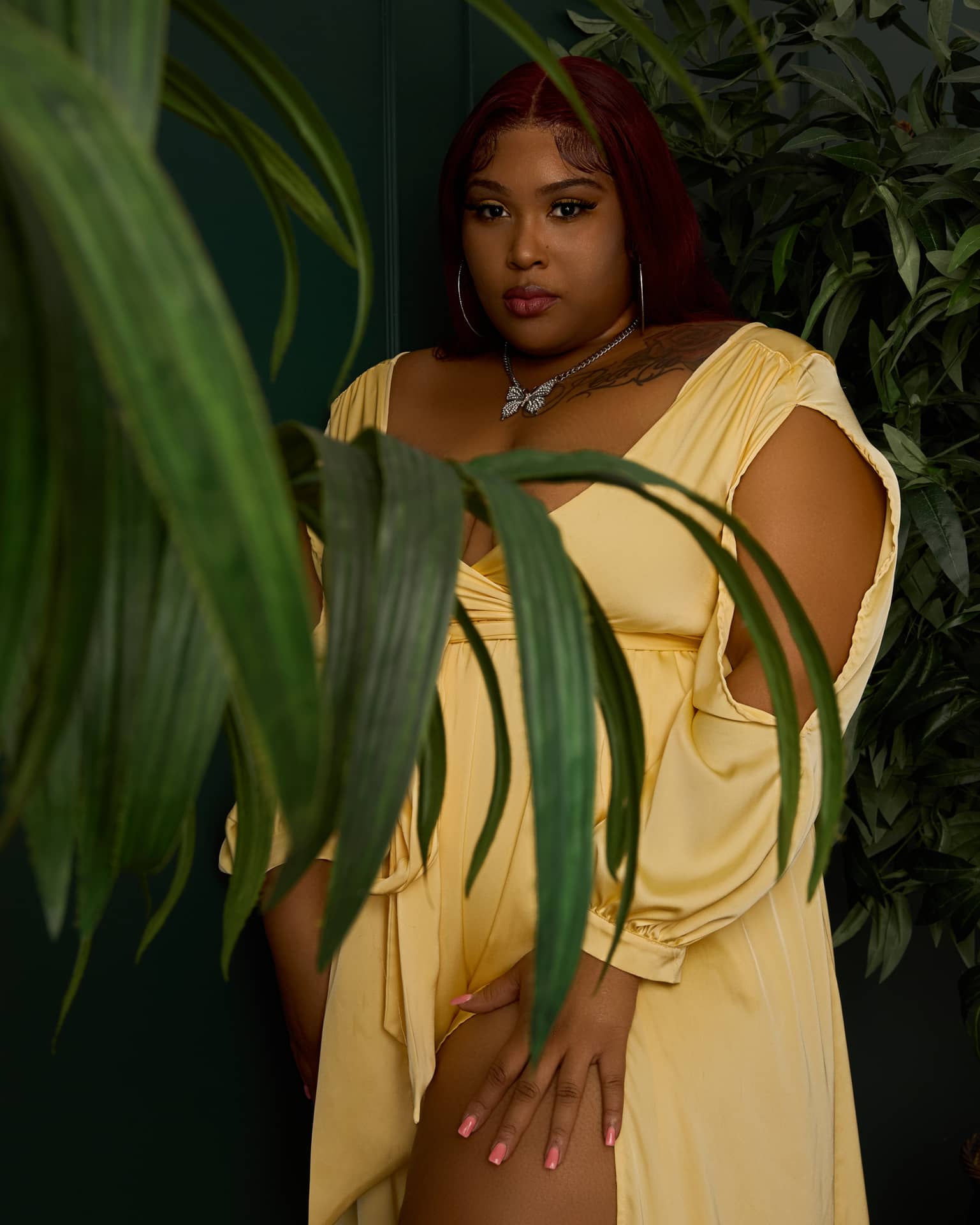Confident woman with red hair poses seated on a wooden stool, wearing a flowing yellow dress with cutout sleeves and clear heels. She smiles warmly, showcasing her tattoos and relaxed body language in a bright studio setting.