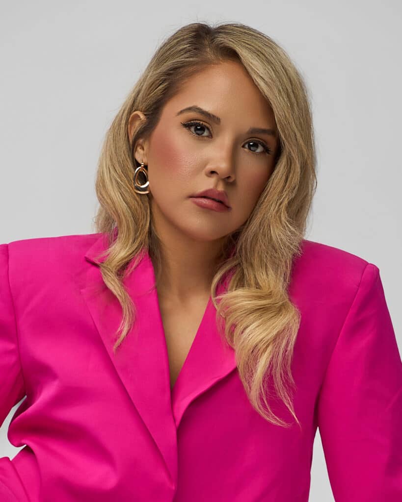 Striking portrait of a poised woman in a vivid fuchsia blazer, styled with gold statement earrings and soft glam makeup. Her direct gaze and confident posture embody quiet strength and modern elegance.