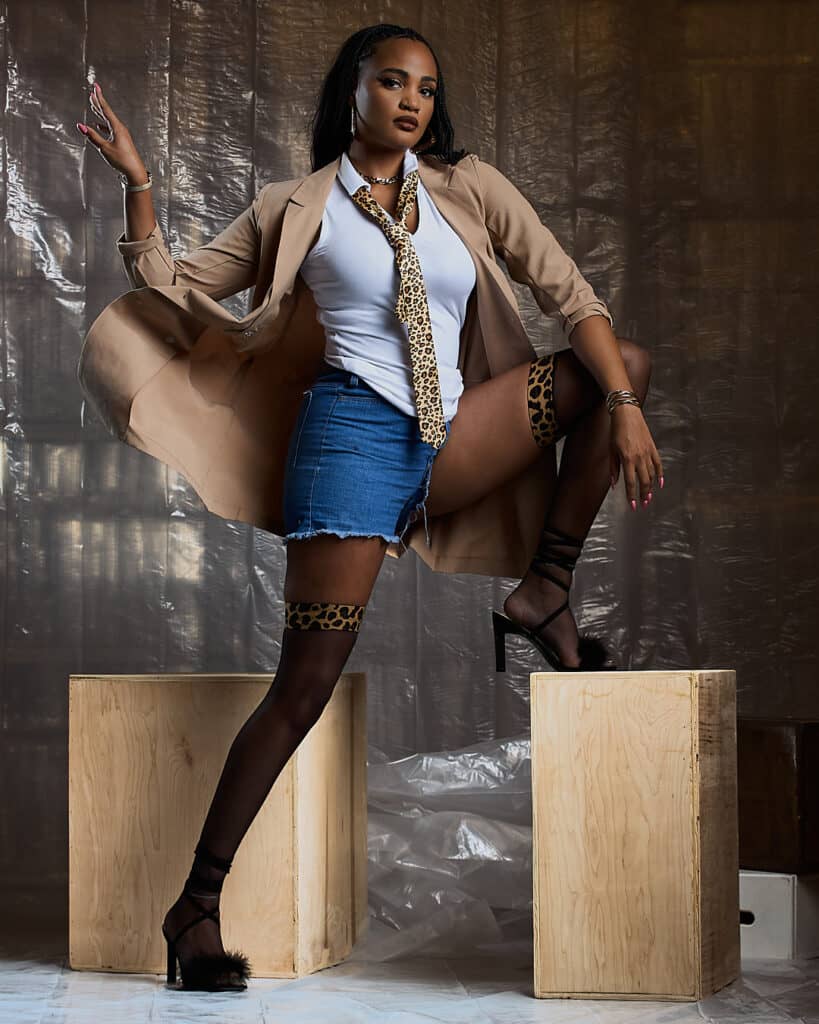An edgy fashion portrait of a woman confidently pulling at a leopard print tie, styled with a beige trench coat, white cutout top, and denim shorts. Leopard print thigh-high stockings and gold accessories complete the look, blending attitude with chic editorial styling.