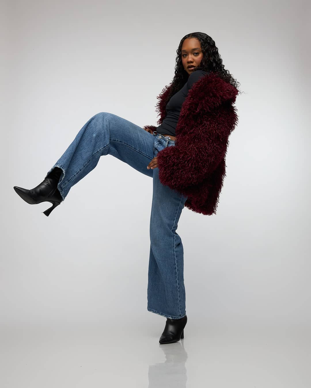 Woman wearing a deep burgundy shag coat and black top, posing confidently against a gray studio background.