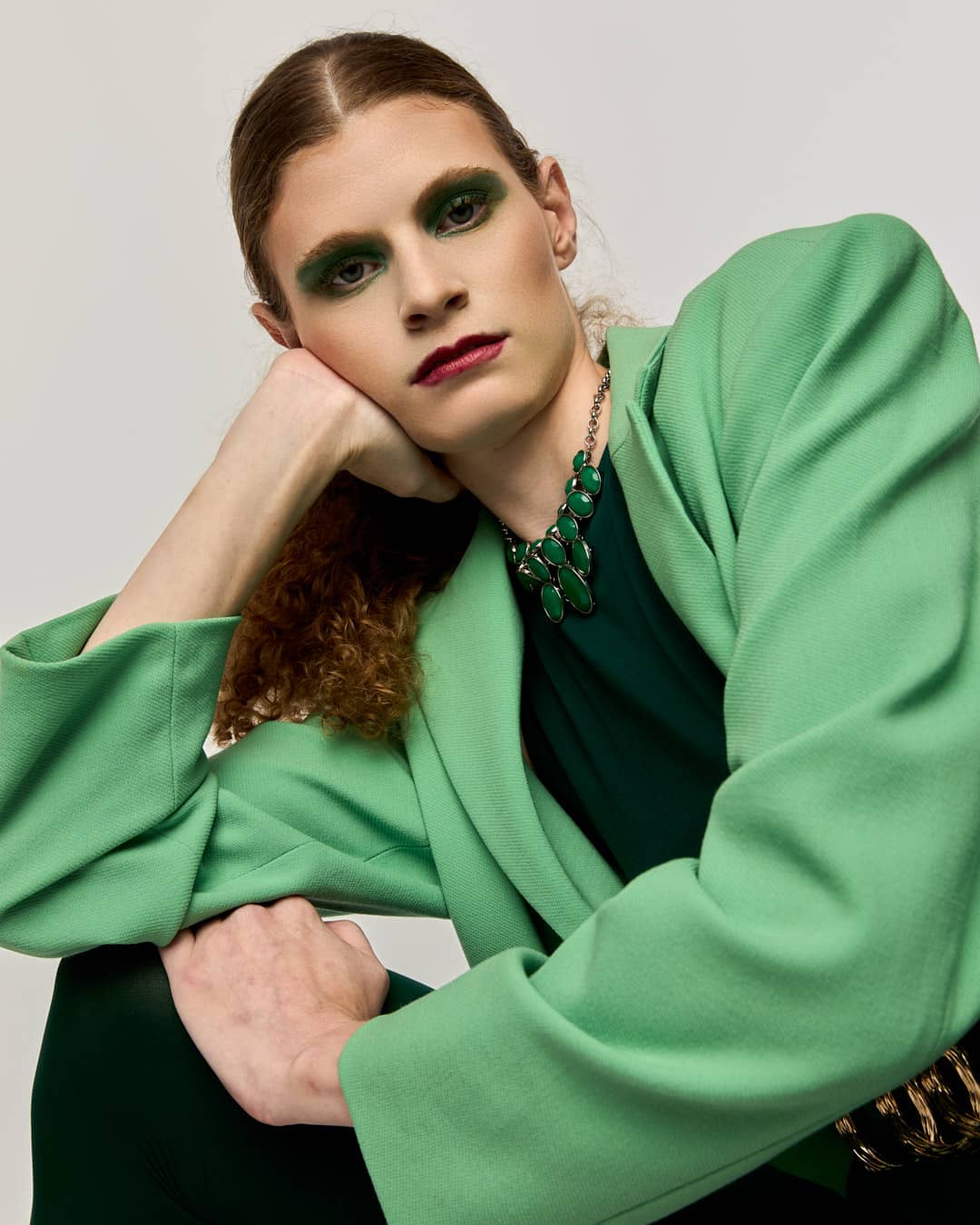 Green-on-green editorial fashion portrait of a model in an oversized mint-green blazer with black tights and green stiletto heels on a light gray studio backdrop.