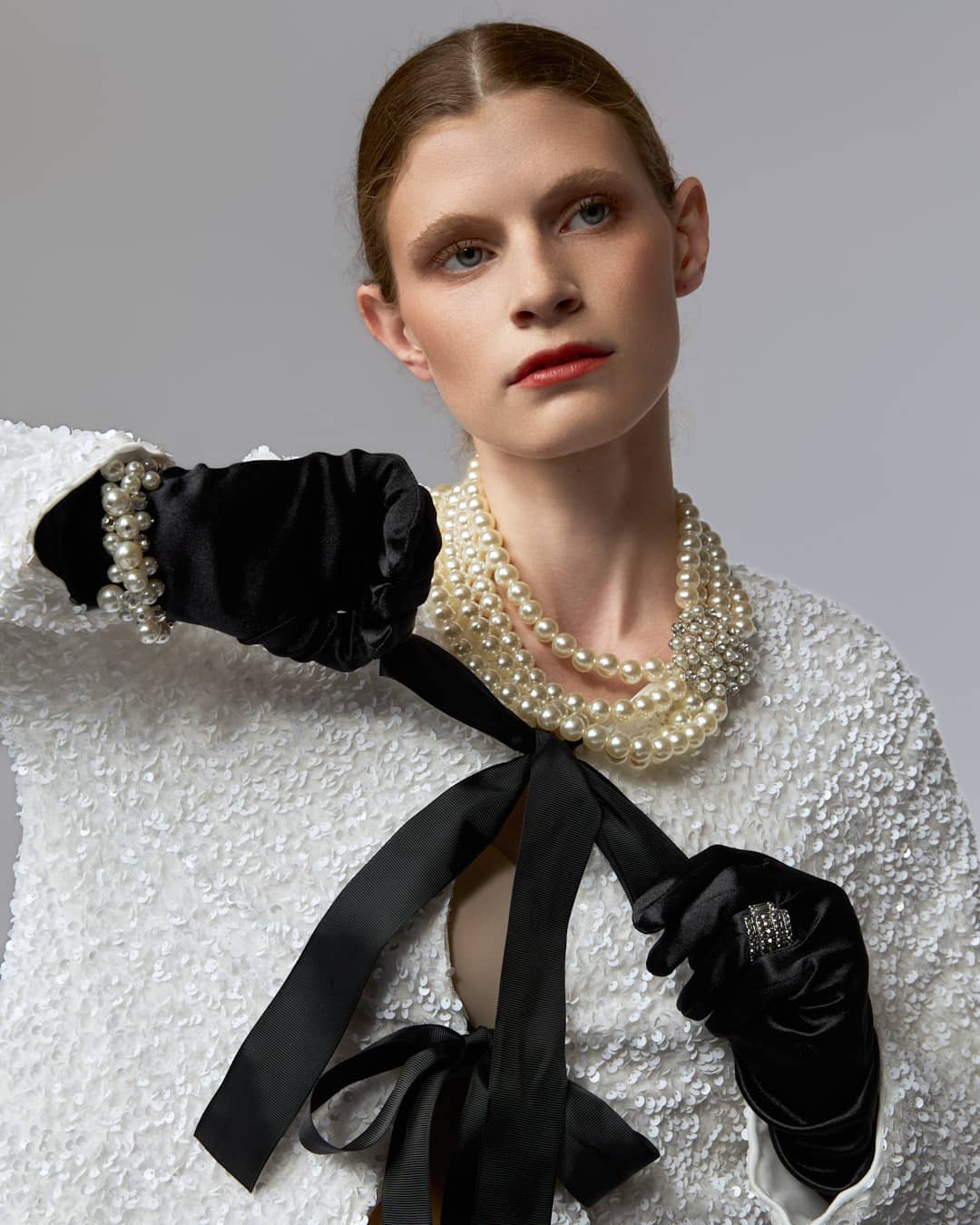 Fashion editorial on a clean gray seamless. The look channels Karl-era tailoring: a cropped white textured jacket finished with oversized black bows, layered pearls, black gloves, wide-leg trousers, and pointed black heels. Minimal set, sharp styling, and a controlled pose keep it graphic and high fashion.