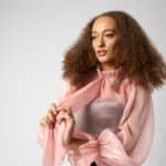 studio fashion portrait of a model in a sheer blush-pink bow blouse with a black midi skirt, wide belt, and black platform heels on a white seamless backdrop.
