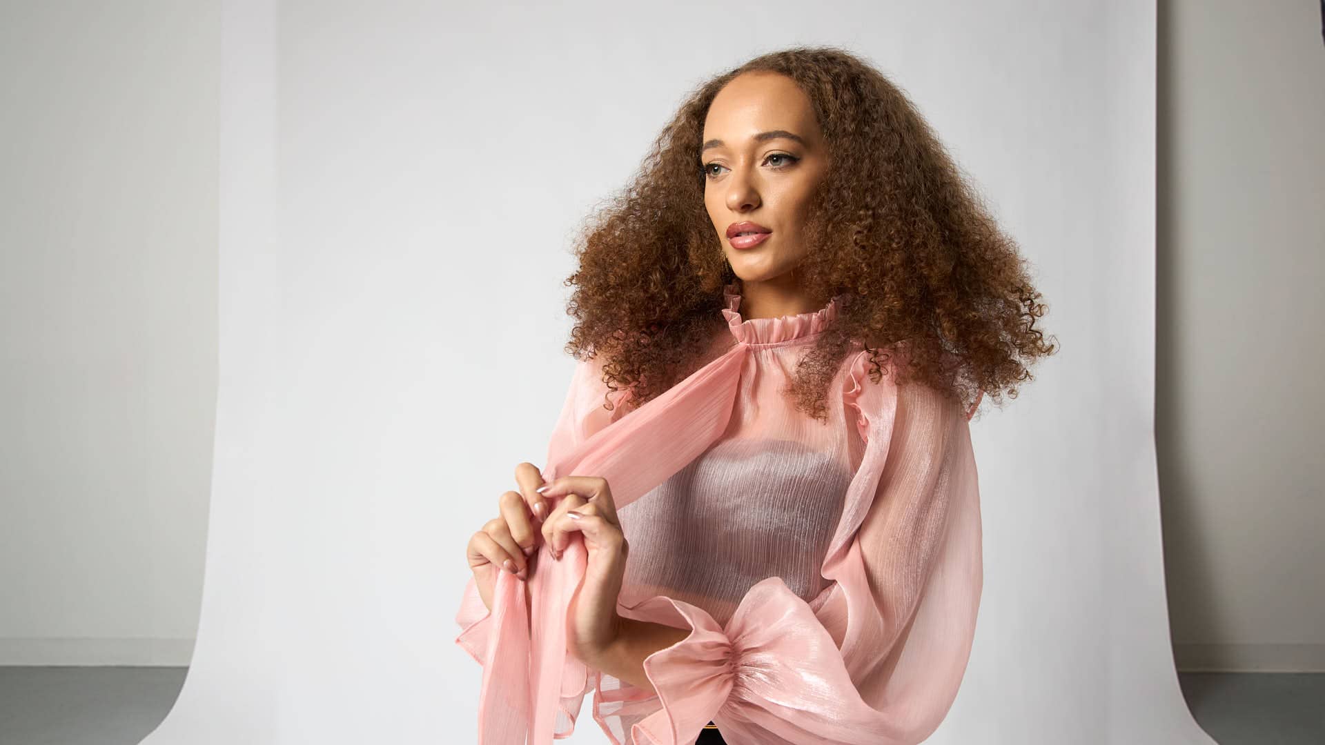 studio fashion portrait of a model in a sheer blush-pink bow blouse with a black midi skirt, wide belt, and black platform heels on a white seamless backdrop.