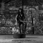 Woman sitting on the floor holding an electric guitar vertically with her back to the camera in front of a colorful graffiti-covered wall.