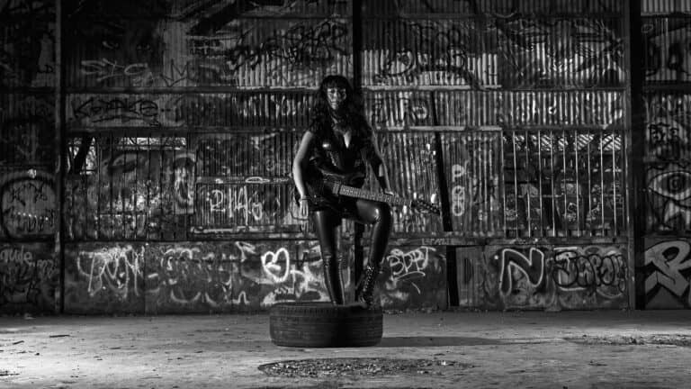 Woman sitting on the floor holding an electric guitar vertically with her back to the camera in front of a colorful graffiti-covered wall.