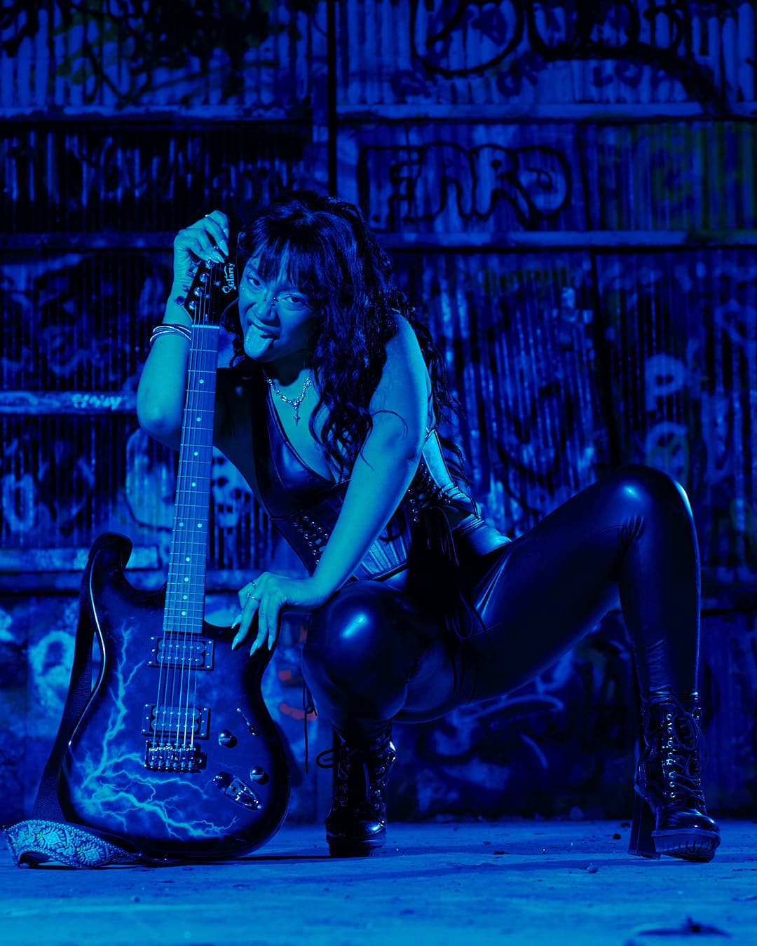 Woman posing in a black leather corset and pants under blue dramatic lighting against a graffiti-covered metal wall.